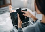 Young Asian woman lying on bed using smartphone and working with laptop on her lap, working from home in the morning